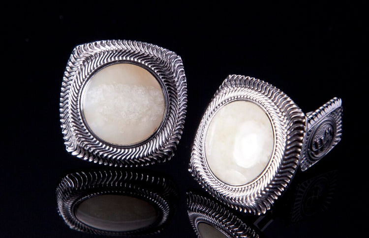 Pair of silver cufflinks with fossil ivory centers, featuring a wavy textured border, reflected on a black surface.