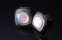 Pair of pearl cufflinks with silver-toned metal frames featuring a wavy etched pattern, displayed on a reflective dark surface.