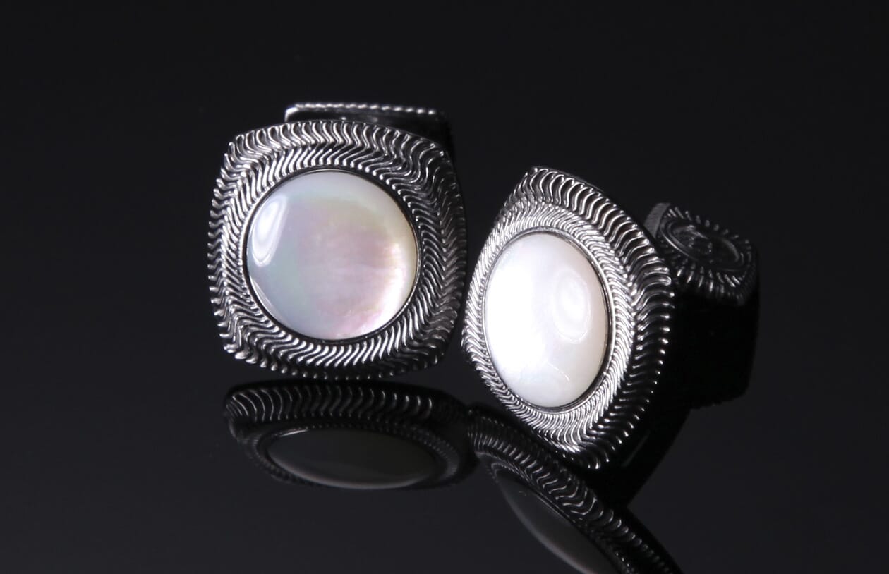 Pair of pearl cufflinks with silver-toned metal frames featuring a wavy etched pattern, displayed on a reflective dark surface.