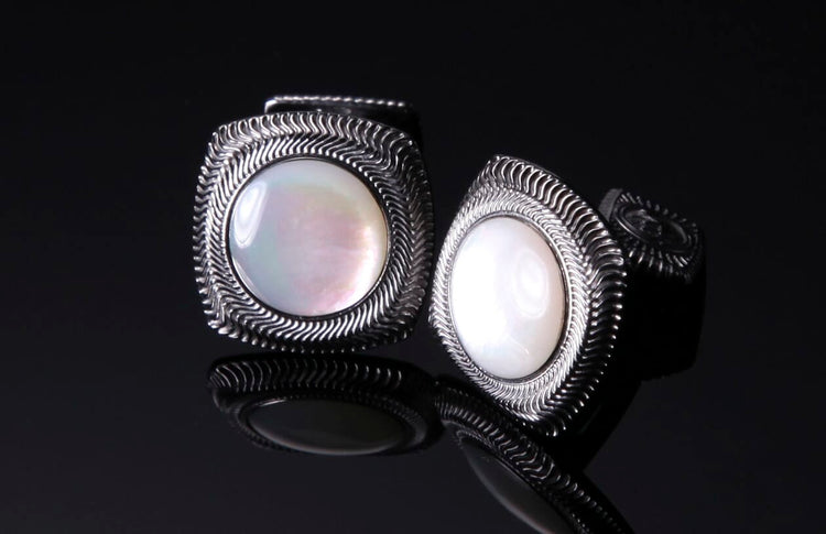 Pair of pearl cufflinks with silver-toned metal frames featuring a wavy etched pattern, displayed on a reflective dark surface.