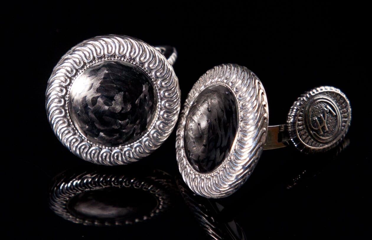 Carbon fiber cufflinks with hammered centers and ornate silver-toned swirling frames, reflecting on a dark surface.