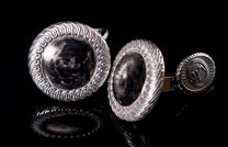 Carbon fiber cufflinks with hammered centers and ornate silver-toned swirling frames, reflecting on a dark surface.