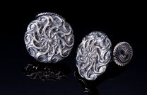 Two metal cufflinks featuring intricate swirling mandala patterns, displayed on a reflective black surface.