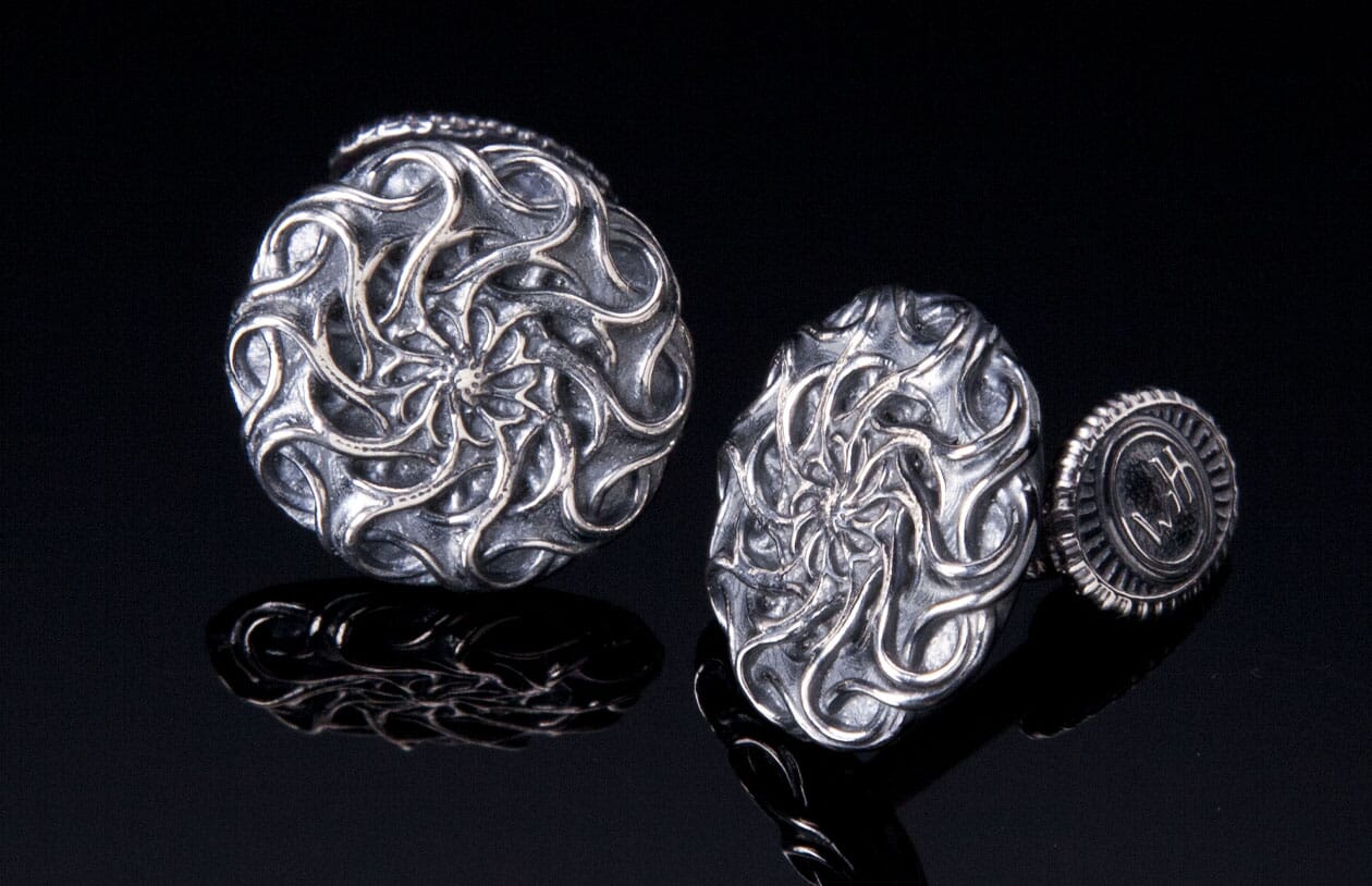 Two metal cufflinks featuring intricate swirling mandala patterns, displayed on a reflective black surface.