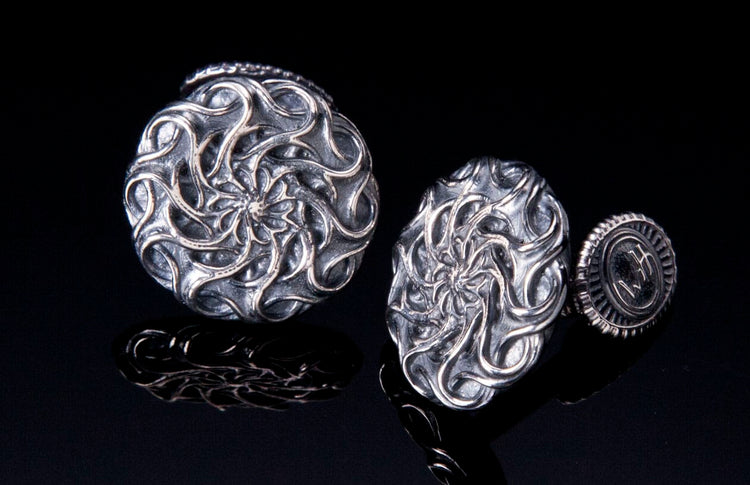 Two metal cufflinks featuring intricate swirling mandala patterns, displayed on a reflective black surface.