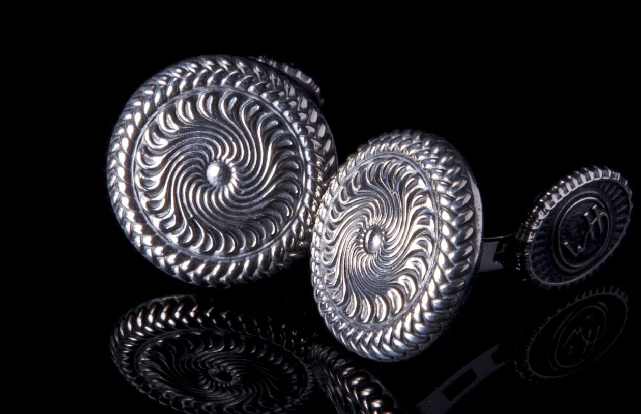 Silver metal cufflinks with intricate swirling patterns and braided edges, reflecting on a dark surface.