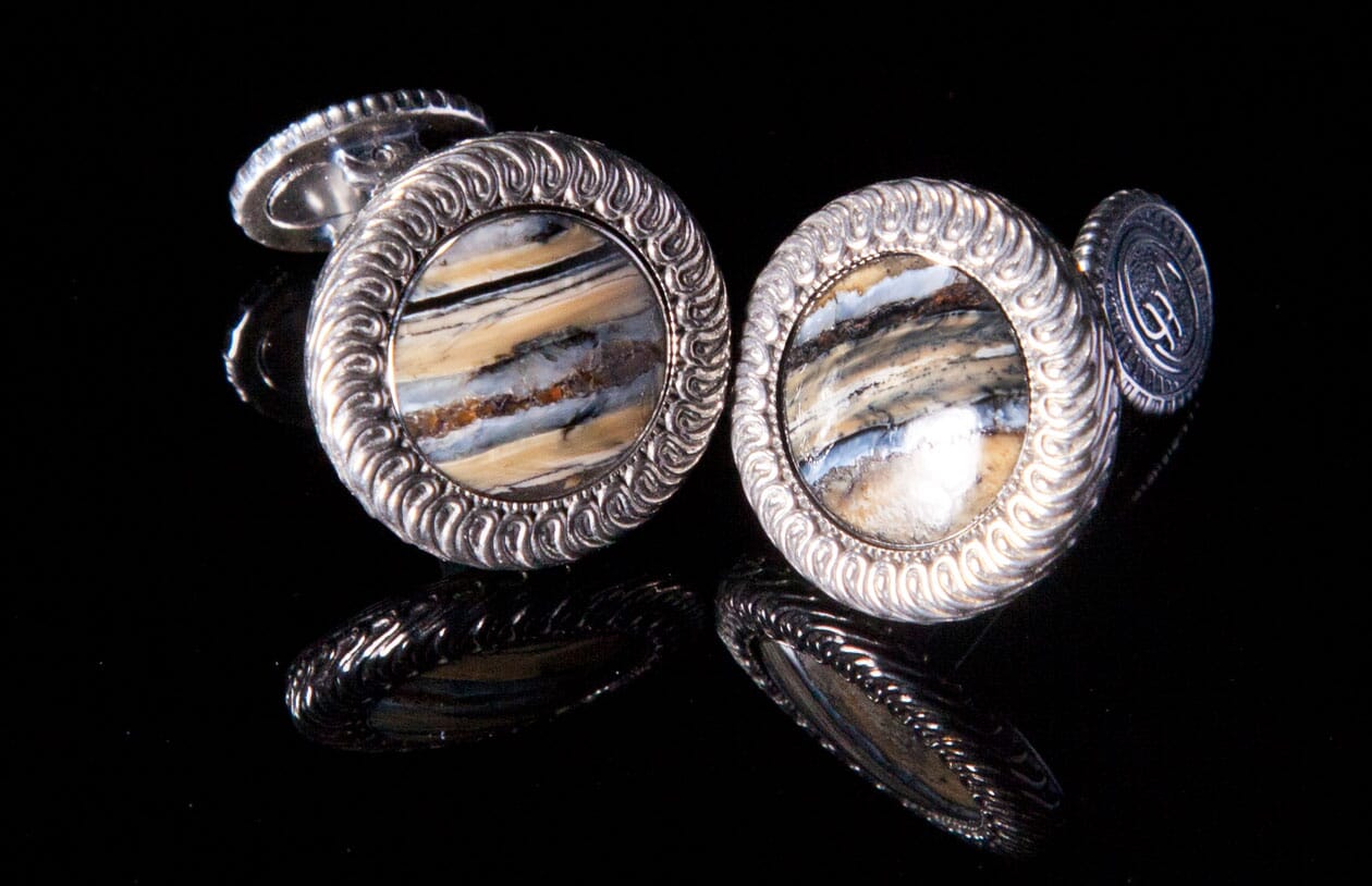 Pair of silver cufflinks with striped mammoth inlay, featuring a scrolled border and reflected on black.