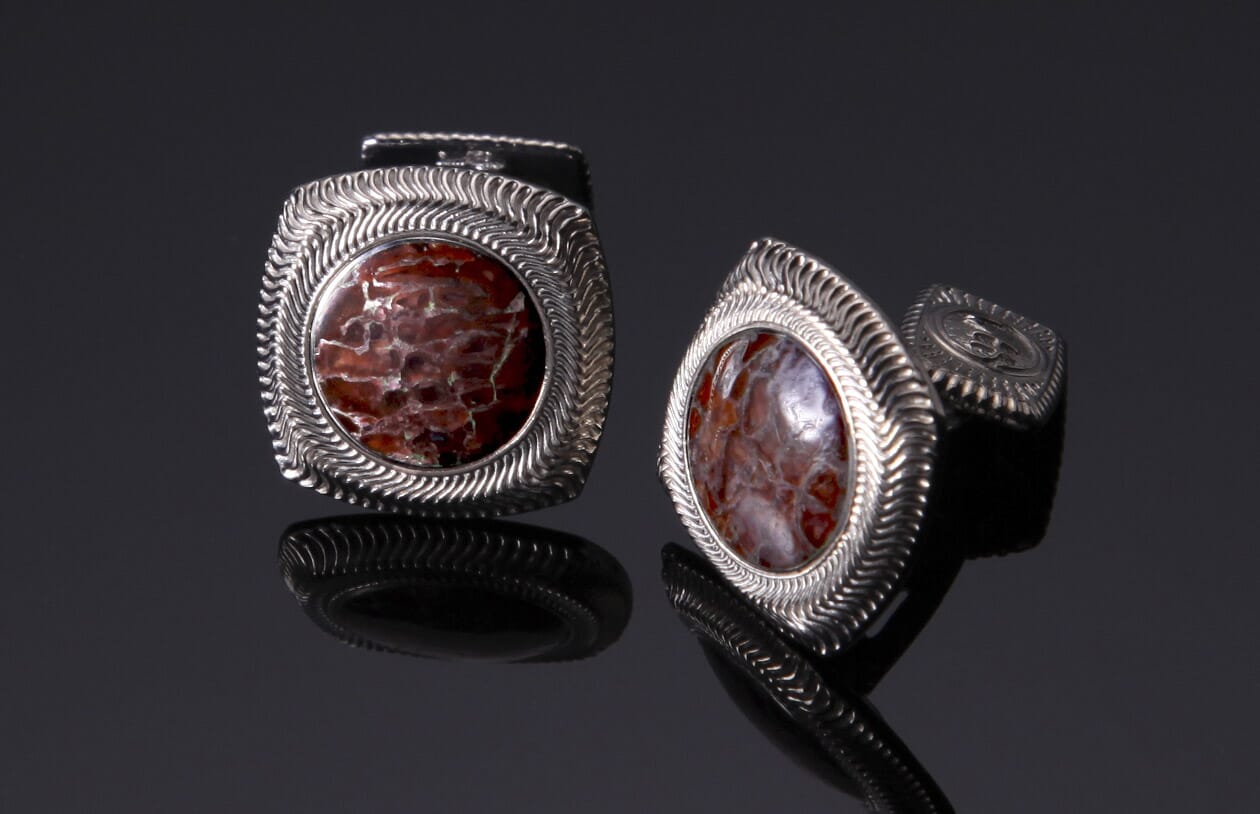 Silver cufflinks with reddish-brown fossilized stone inserts and wavy textured frames, displayed on a reflective surface.