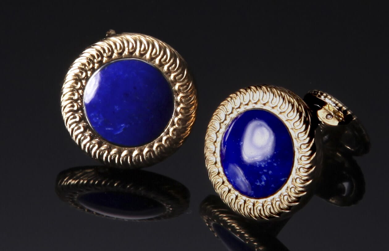 Lapis lazuli cufflinks with ornate gold bezels, reflecting on a dark surface.