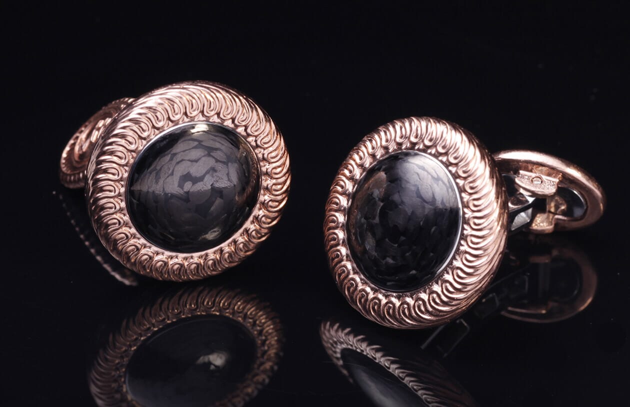 Pair of rose gold cufflinks with engraved wavy borders and black carbon fiber oval centers, displayed with reflections.