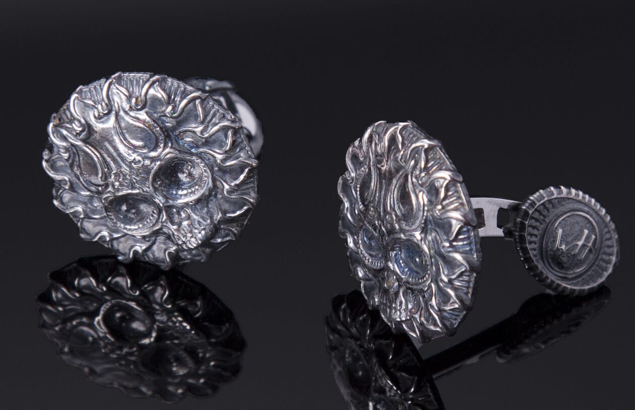 A pair of silver skull cufflinks with intricate flaming designs, blue eye accents, and a back mechanism showing 'WH' initials.