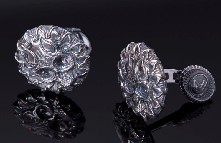 A pair of silver skull cufflinks with intricate flaming designs, blue eye accents, and a back mechanism showing 'WH' initials.