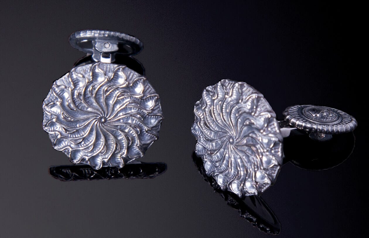 Silver-toned cufflinks featuring a swirling leaf pattern, one upright and the other on its side revealing a circular etched back.