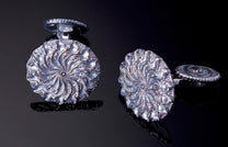 Silver-toned cufflinks featuring a swirling leaf pattern, one upright and the other on its side revealing a circular etched back.
