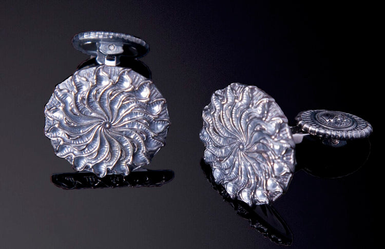 Silver-toned cufflinks featuring a swirling leaf pattern, one upright and the other on its side revealing a circular etched back.