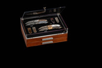 Wooden collector's box with an open clear acrylic top, displaying ornate knives and money clips on a black interior, over two drawers with silver handles.
