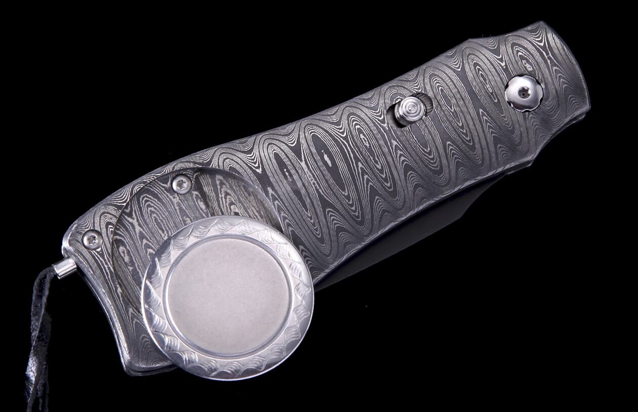 Folding knife with a swirling Damascus steel handle, featuring an attached round silver-toned medallion, on a black background.