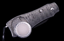 Folding knife with a swirling Damascus steel handle, featuring an attached round silver-toned medallion, on a black background.