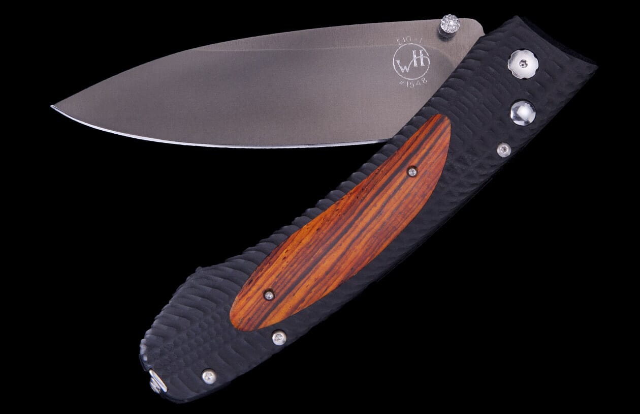 Open folding knife with a dark grey blade, black textured handle, and reddish-brown wood inlay. The blade features an etched 'WH' logo and 'E10-1'.