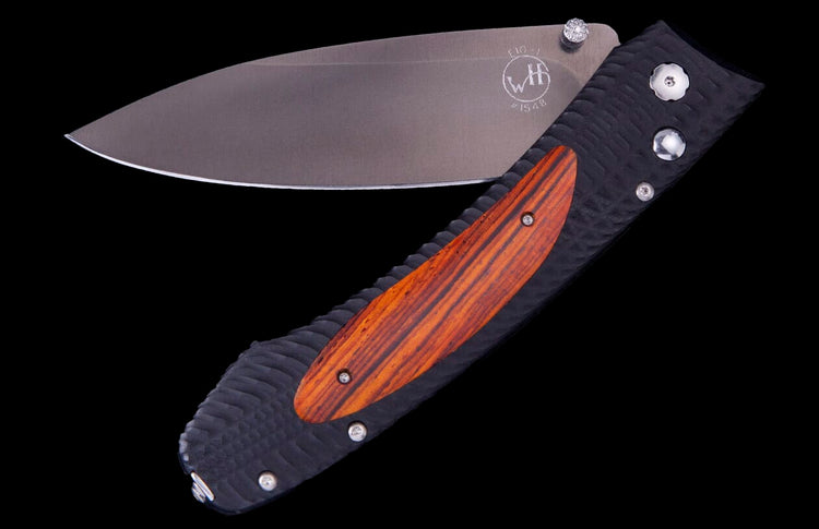 Open folding knife with a dark grey blade, black textured handle, and reddish-brown wood inlay. The blade features an etched 'WH' logo and 'E10-1'.