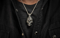 Ornate silver skull pendant with blue gemstones on a textured chain, worn over a black ribbed shirt and denim jacket.