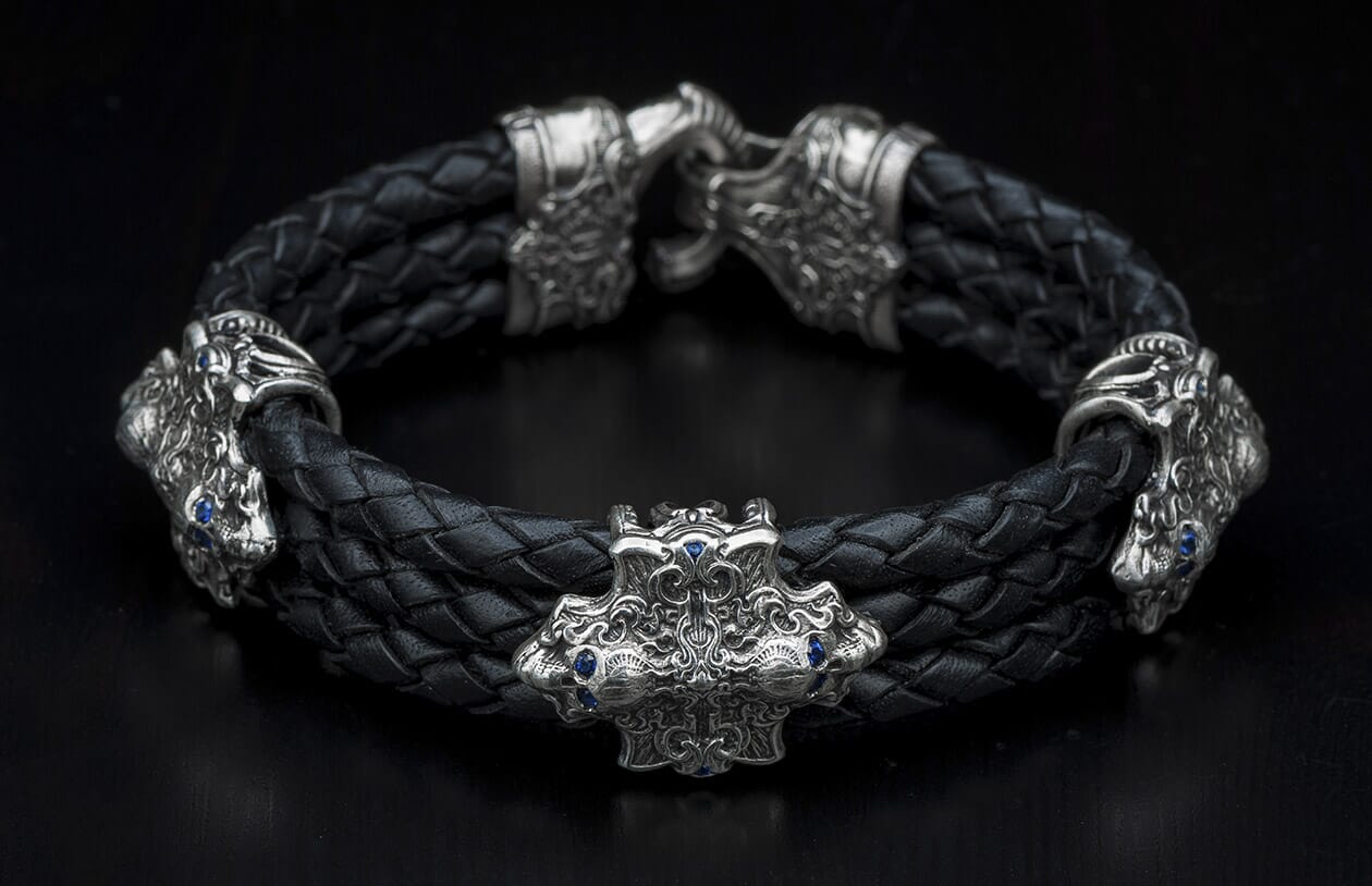 Black braided leather bracelet with silver accents depicting ornate creature heads and a central crest adorned with blue gemstones.