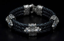 Black braided leather bracelet with silver accents depicting ornate creature heads and a central crest adorned with blue gemstones.