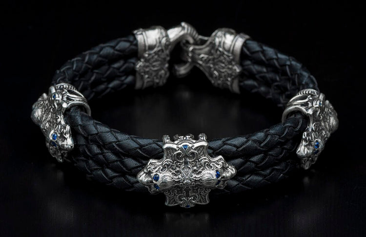 Black braided leather bracelet with silver accents depicting ornate creature heads and a central crest adorned with blue gemstones.
