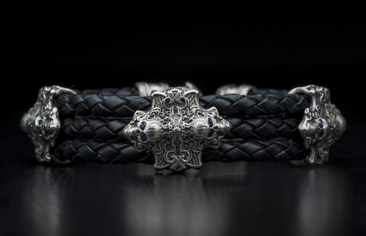 Black braided leather bracelet with an ornate silver-toned cross-shaped centerpiece featuring blue gemstones and skull motifs.