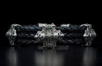 Black braided leather bracelet with an ornate silver-toned cross-shaped centerpiece featuring blue gemstones and skull motifs.