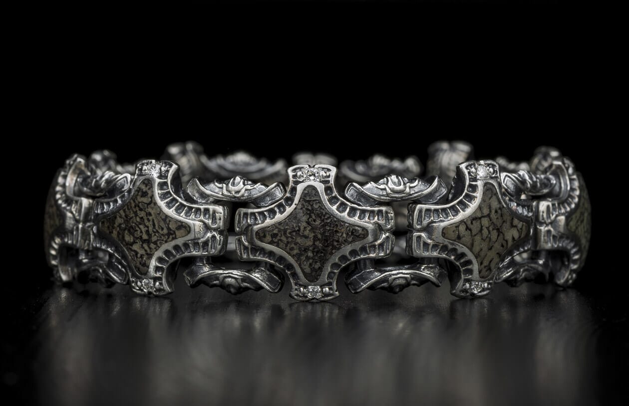 Ornate silver bracelet featuring intricate links, textured dark inserts, and small clear stones, on a reflective black surface.