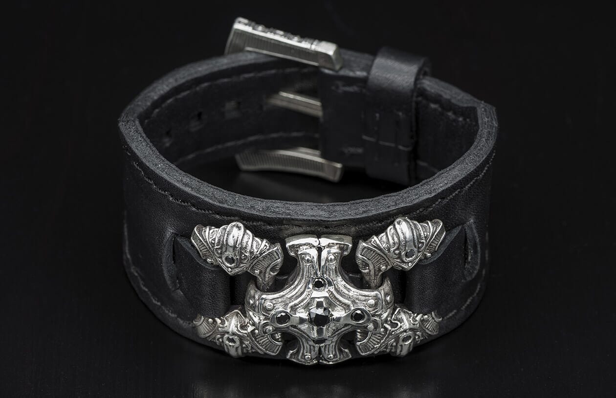 Black leather cuff bracelet with an ornate silver-tone cross-shaped emblem detailed with black stones and a buckle closure.