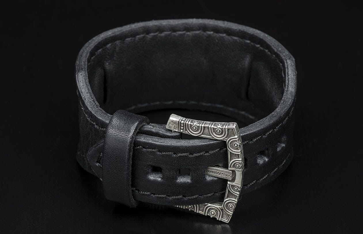Black leather bracelet with a decorative silver-toned buckle featuring intricate patterns.