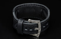 Black leather bracelet with a decorative silver-toned buckle featuring intricate patterns.