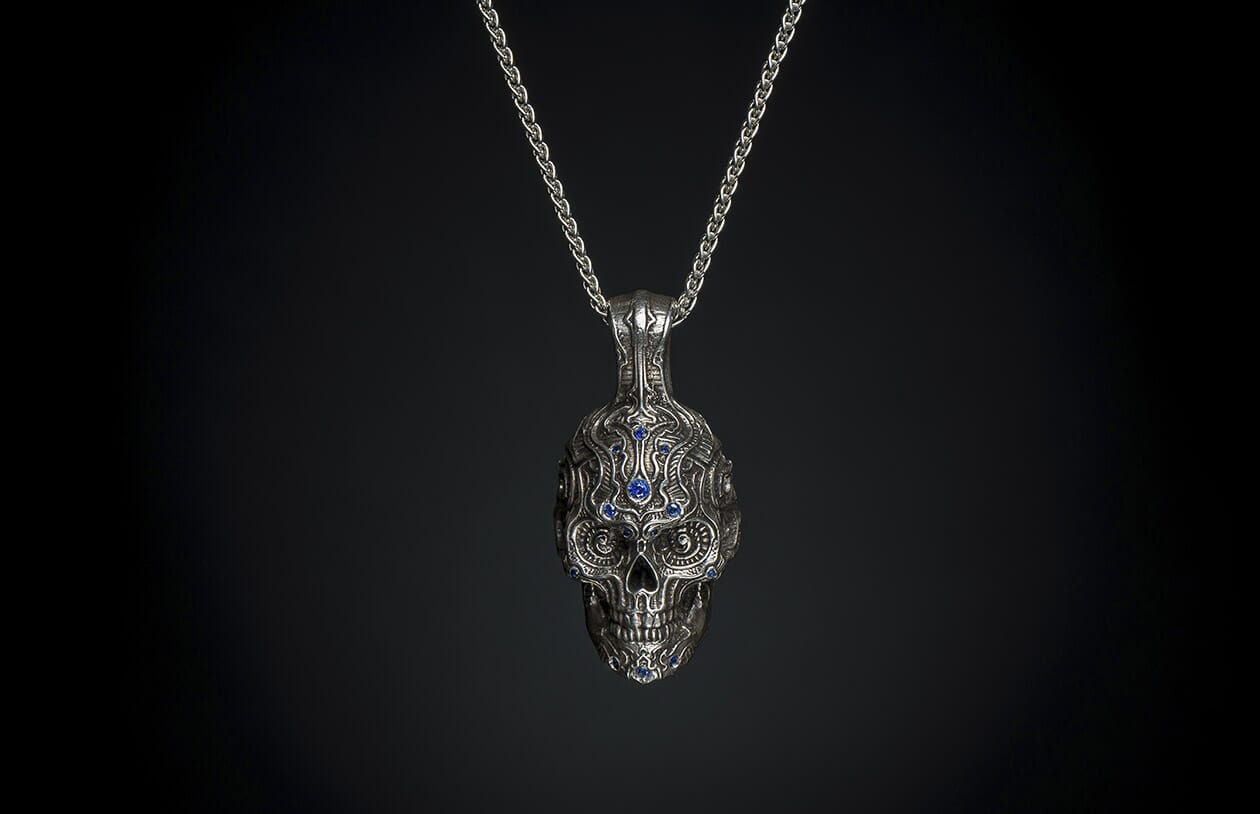 Ornate silver skull pendant with blue gemstones, front view, hanging from a silver chain against a dark background.