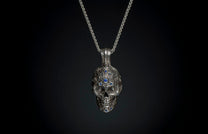 Ornate silver skull pendant with blue gemstones, front view, hanging from a silver chain against a dark background.