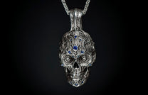 Silver skull pendant with intricate ornate carvings and blue gemstones, hanging from a metal chain.