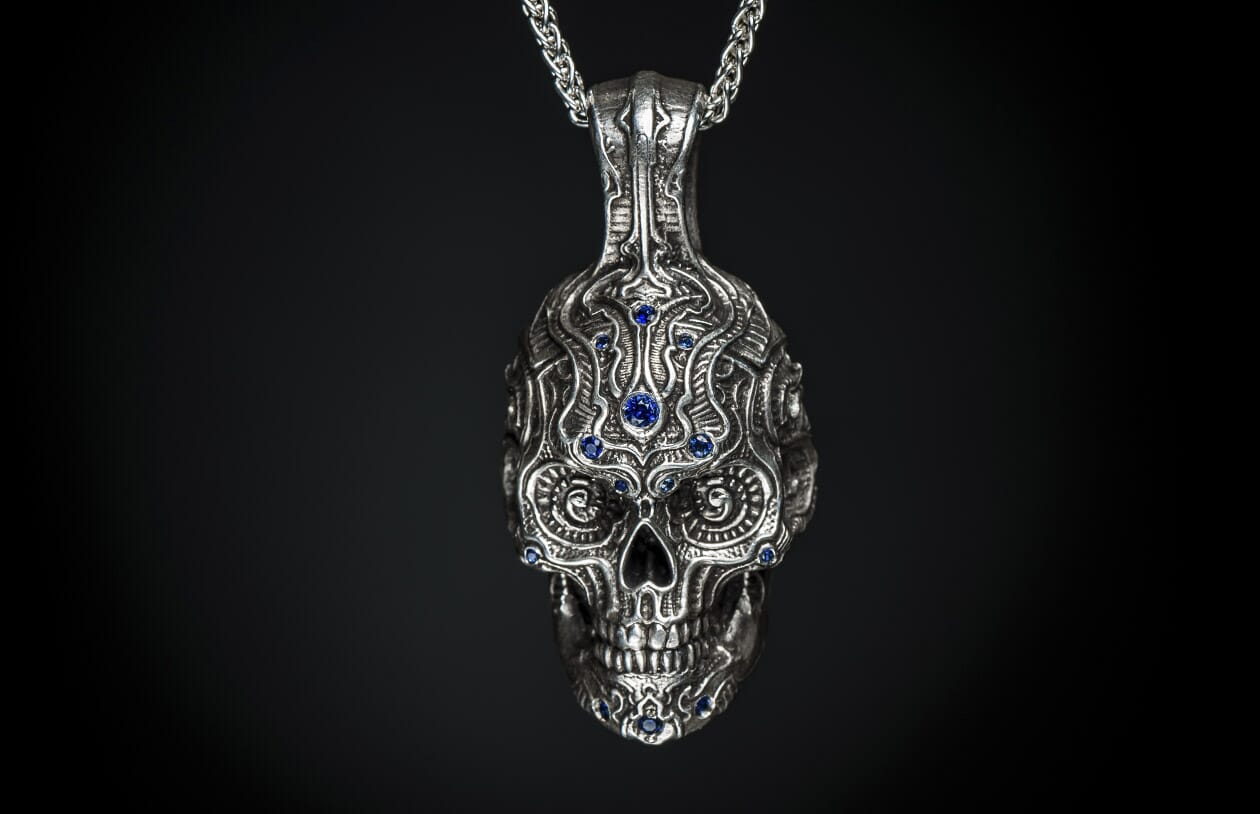 Silver skull pendant with intricate ornate carvings and blue gemstones, hanging from a metal chain.