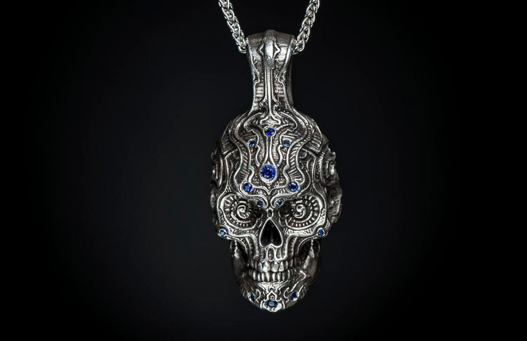 Silver skull pendant with intricate ornate carvings and blue gemstones, hanging from a metal chain.