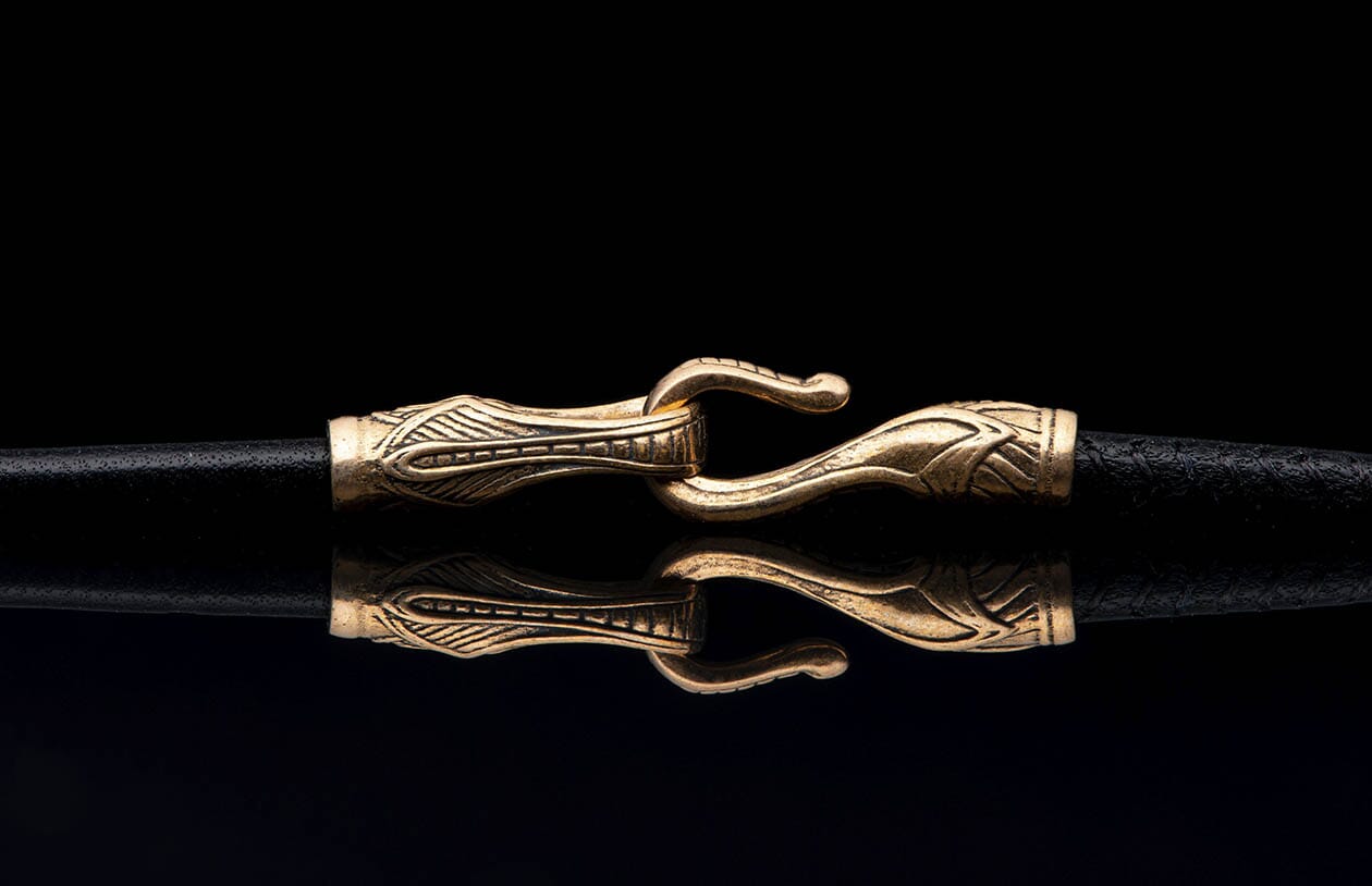 Ornate gold-toned jewelry clasp with intricate patterns securing a black cord, reflected on a dark surface.
