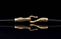 Ornate gold-toned jewelry clasp with intricate patterns securing a black cord, reflected on a dark surface.