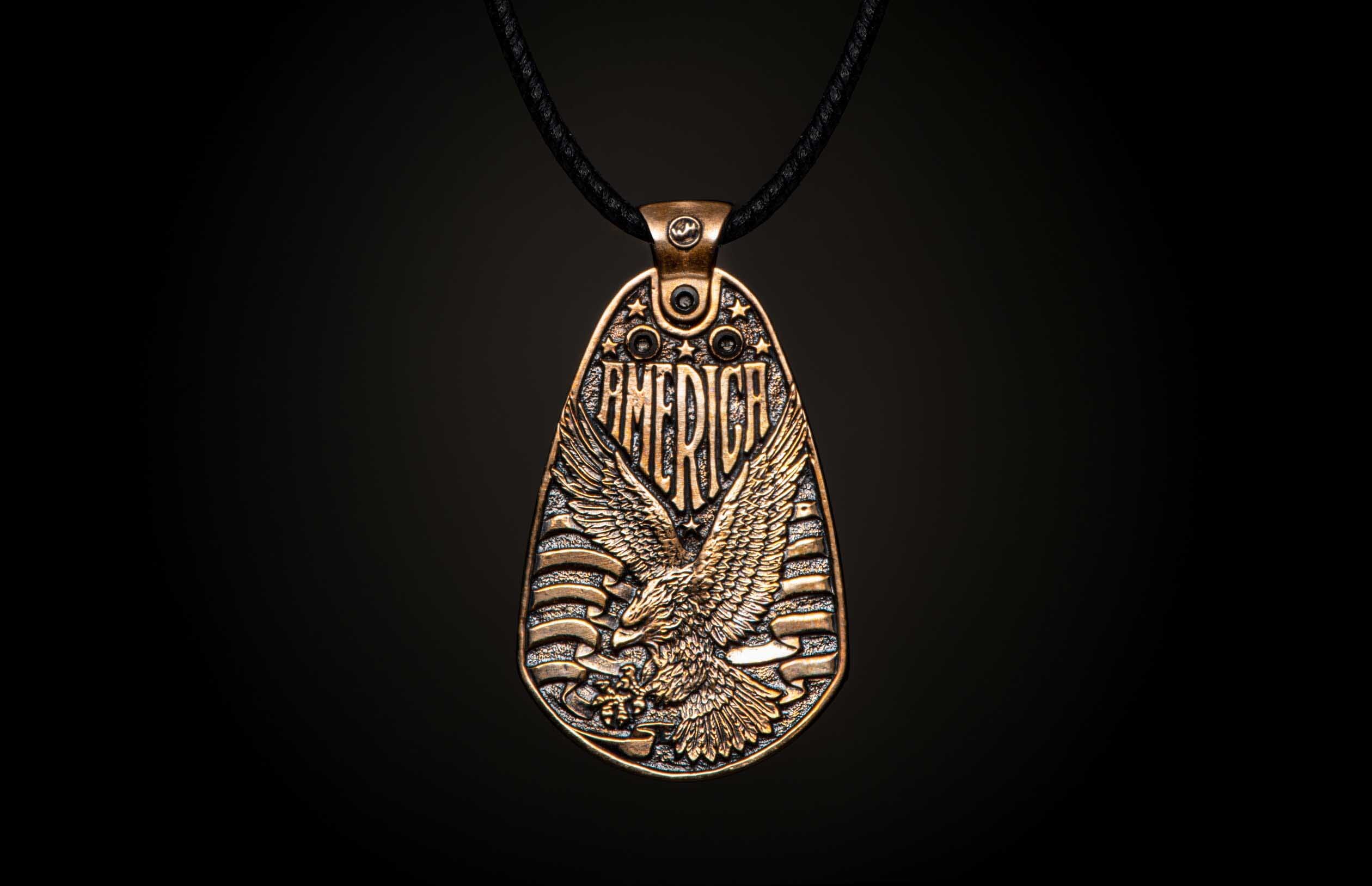 Aged bronze pendant necklace featuring a bald eagle, "AMERICA" text, stars, and stripes, suspended from a black cord.
