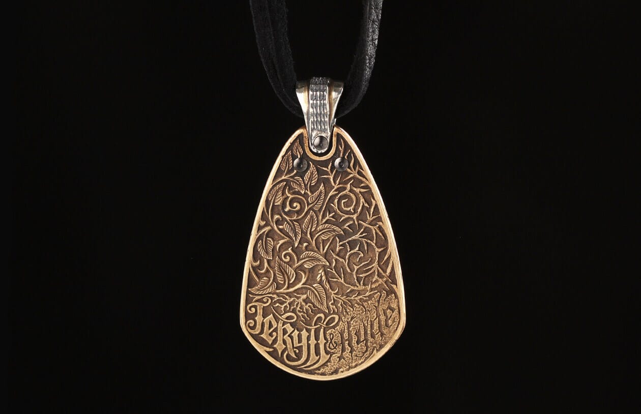 A bronze teardrop pendant by Jekyll + Hyde, featuring intricate leaf and vine carvings with text, hanging from a black cord.
