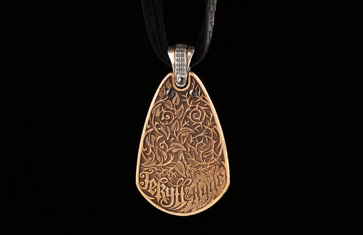 A bronze teardrop pendant by Jekyll + Hyde, featuring intricate leaf and vine carvings with text, hanging from a black cord.