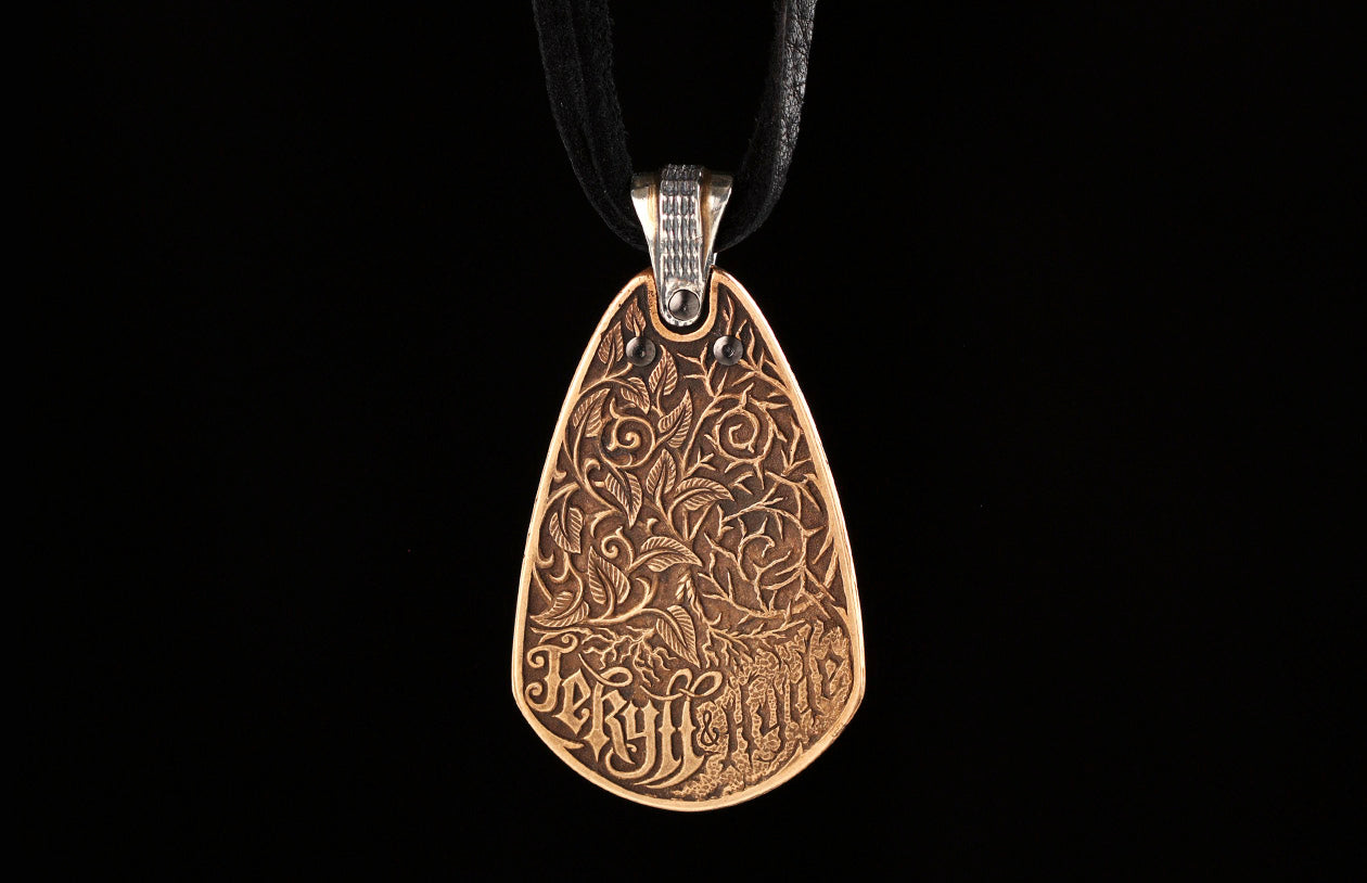 JEKYLL+HYDE-BR - 20"|Bronze pendant with intricate leaf and vine patterns, "Jekyll &amp; Hyde" inscription, hanging from a dark leather cord.