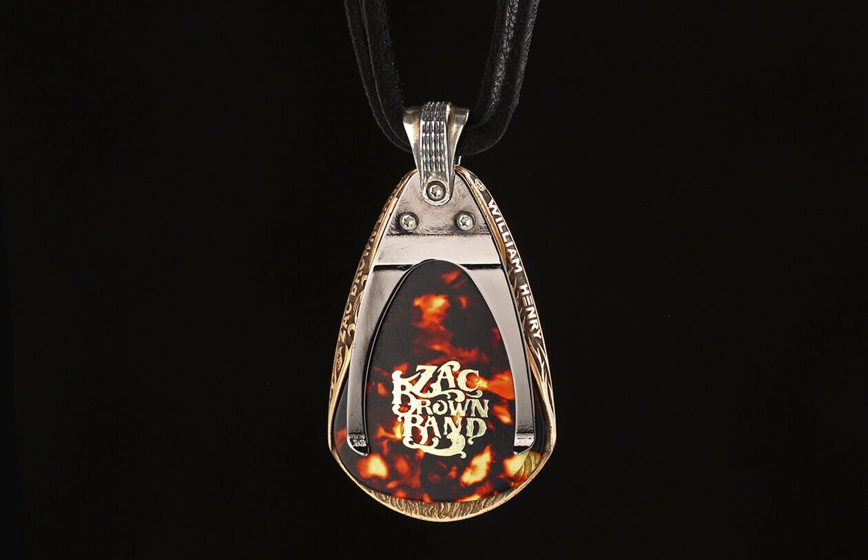 Bronze and silver guitar pick pendant by William Henry with engraved scrollwork, featuring a tortoise-shell pick with 'Zac Brown Band' logo, on a black cord.