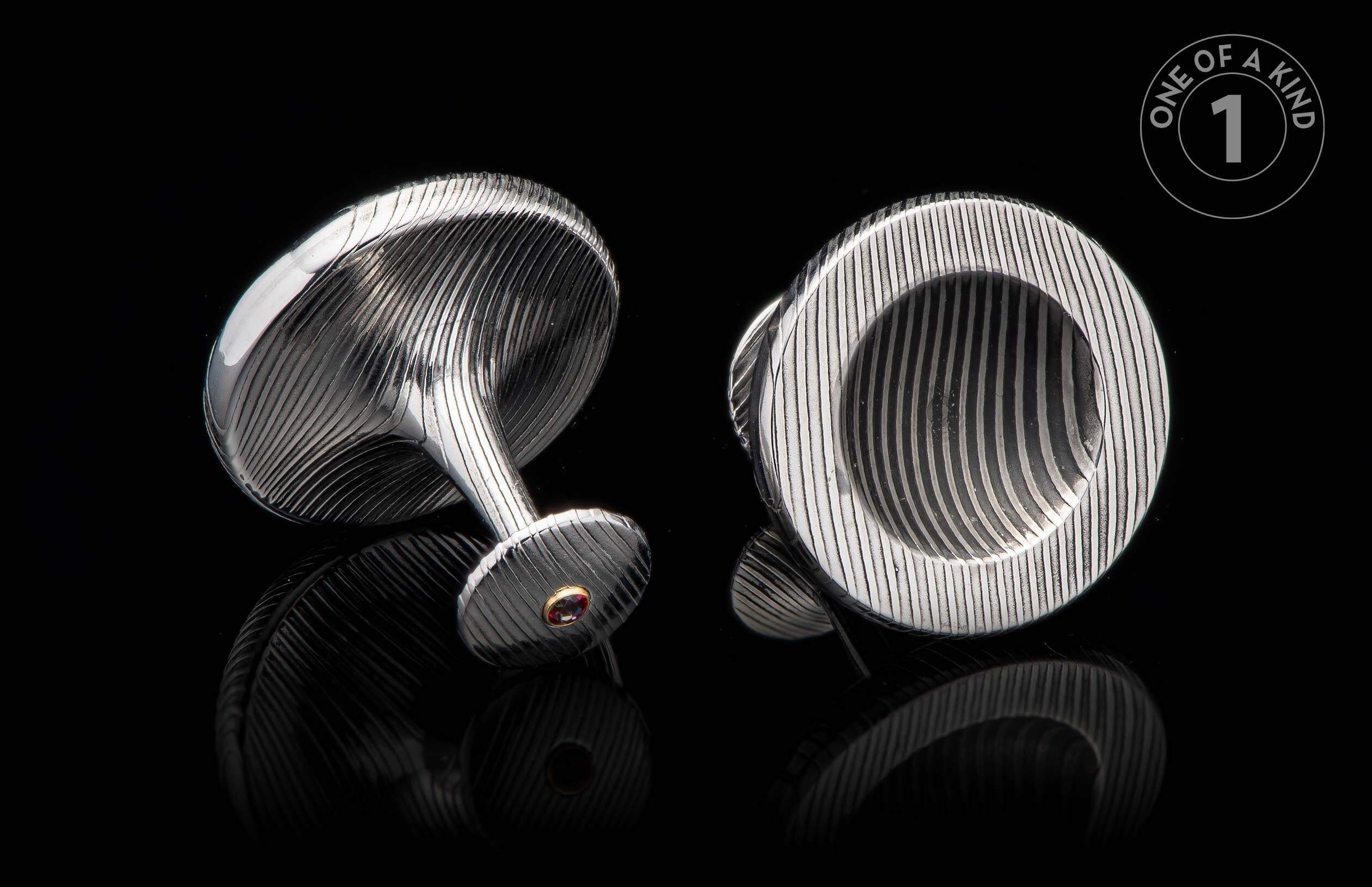 Pair of silver-toned cufflinks with engraved wavy patterns, one featuring a red gemstone in a gold bezel.