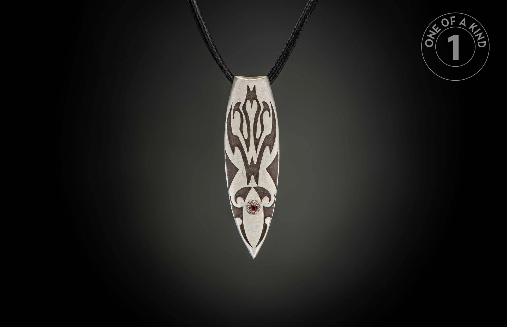 Silver pendant with dark etched patterns, a red gemstone, and black braided cord.
