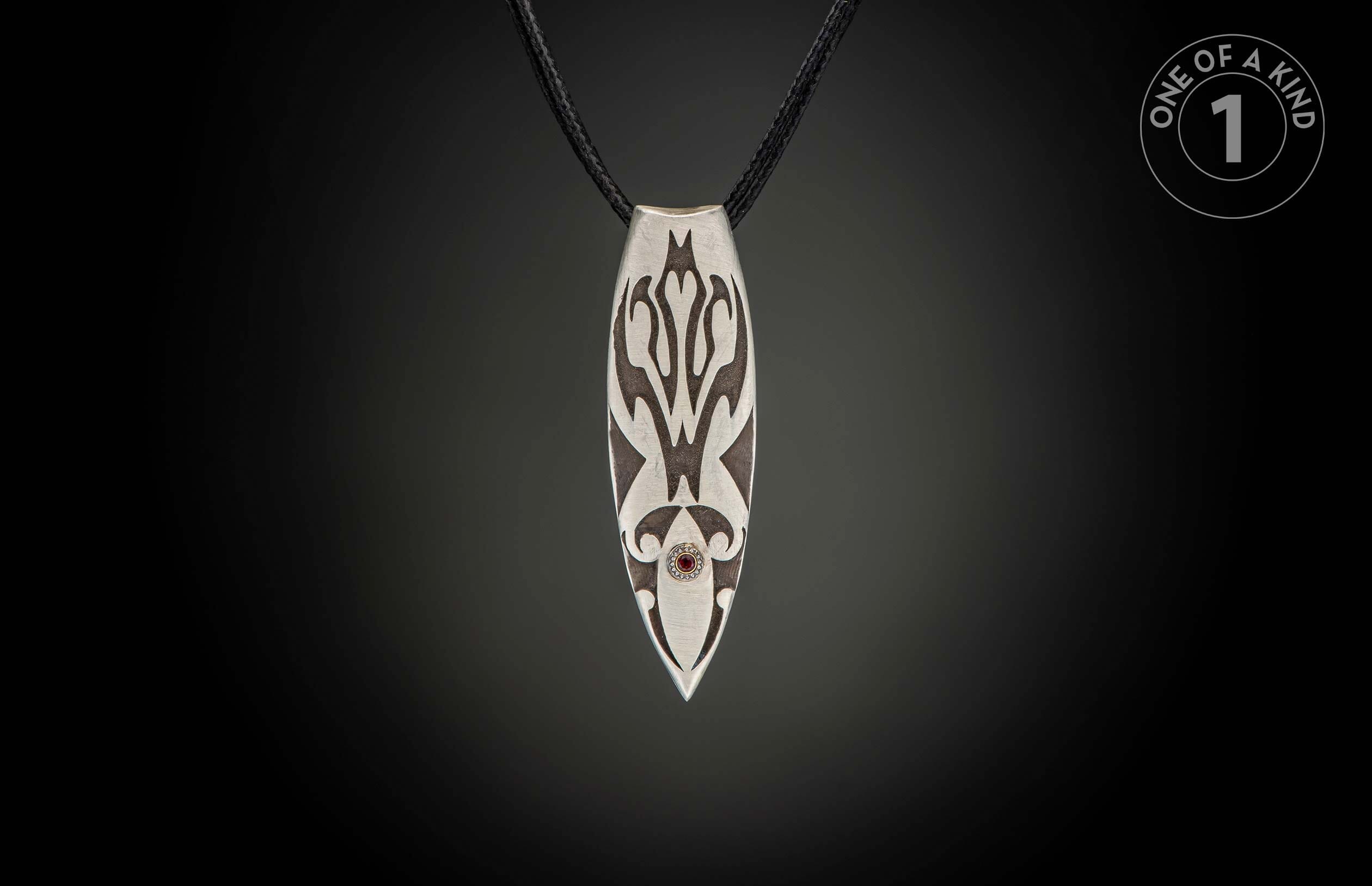 Silver pendant with dark etched patterns, a red gemstone, and black braided cord.