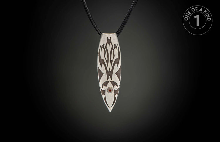 Silver pendant with dark etched patterns, a red gemstone, and black braided cord.